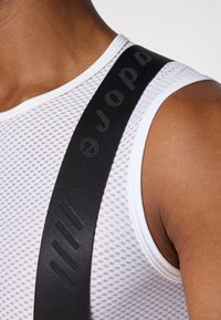 Schwarze elastische Hosenträger mit erhabenem Logo, befestigt an einem weißen Netz-Top mit strukturiertem Muster und einem gerippten Ausschnitt.