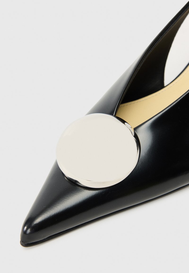 Zapato de charol negro con punta afilada que presenta un acento circular plateado en la parte superior. Interior tapizado con material beige.