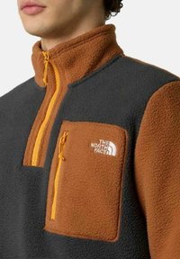 Sudadera de polar en gris oscuro y marrón, con un bolsillo en el pecho con cremallera, cuello alto y detalles en naranja en la cremallera y el bolsillo.