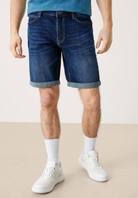 Tummat farkkushortsit, joissa on käännetyt lahkeet, viisi taskua ja klassinen malli. Yhdistettynä valkoisiin tennareihin ja ribattuvalkoisiin sukkiin.