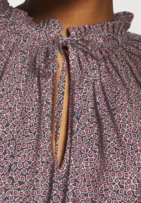 Blouse en tissu sombre avec un motif géométrique rouge et blanc, dotée d'un col volants et d'une fermeture à nouer à l'avant.