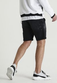 Pantalones cortos de mezclilla negra con dobladillos enrollados, que presentan un aspecto desgastado. Se llevan con zapatillas deportivas en blanco y negro. El fondo es neutro.