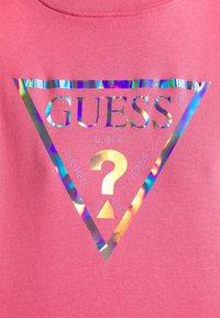 Logo holographique de Guess avec un grand point d'interrogation à l'intérieur d'un triangle sur un fond en tissu rose.