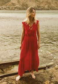 Red, maxi-length dress with ruffled sleeves and a waist tie. Made of lightweight fabric, featuring a tiered skirt. Model stands barefoot by water.