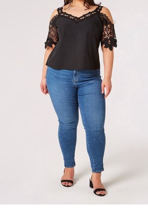 Femme portant un haut noir en dentelle à épaules dénudées, un jean skinny bleu, et des talons ouverts noirs, debout sur un fond blanc.