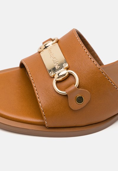 Braune Leder-Sandale mit goldfarbener Hardware-Beschichtung, ausgestattet mit einem rechteckigen Namensschild und einem kreisförmigen Ringdetail. Flaches Sohlen-Design.