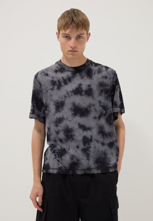 Junger Mann mit schwarz-grauem Batik-Kurzarm-T-Shirt und schwarzen Cargohosen steht vor einem schlichten hellen Hintergrund.