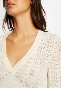 Hellecremefarbene Strickjacke mit V-Ausschnitt, aufwendigem Zickzackmuster und gerippten Akzenten. Weiche Textur und figuraffine Silhouette.