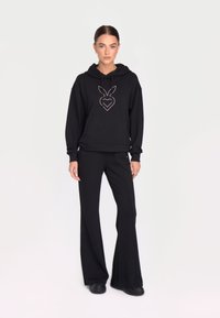 Sweatshirt negru cu glugă și un desen ușor conturat cu un cerb pe față, asortat cu pantaloni negri largi și încălțăminte întunecată.