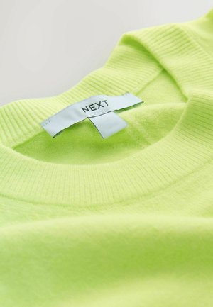 Neonvihreä neulepusero, joka on valmistettu pehmeästä kankaasta. Siinä on resorikaulus ja ommeltu etiketti, jossa lukee mustalla tekstillä "NEXT".