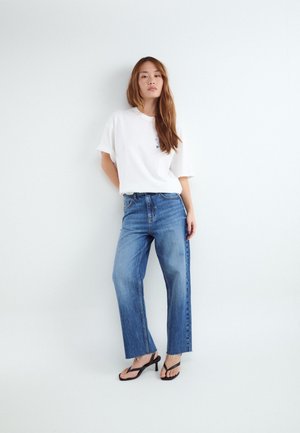 Kvinde med langt hår iført en hvid t-shirt, blå vidde bukser og sorte hælede sandaler, der står op ad en hvid væg.
