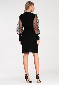 Czarna sukienka z przezroczystymi, kropkowanymi bufkami, dopasowanym gorsetem i lekko rozkloszowanym dołem. Posiada czarne mankiety z złotymi guzikami.