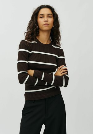 Femme aux cheveux bouclés portant un pull côtelé marron foncé avec des rayures blanches et un pantalon noir, debout les bras croisés devant un fond uni.