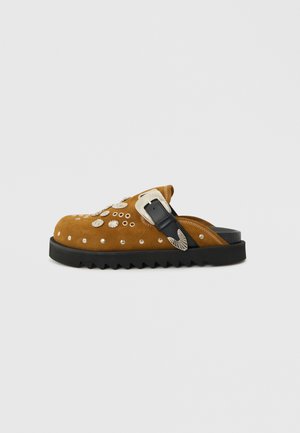 Hnedý semišový slip-on šľapák so striebornými cvočkami a ozdobnou prackou, s hrubou čiernou ryhovanou podrážkou.