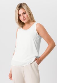 Haut sans manches blanc avec un col rond, texture lisse et subtile brillance ; associé à un pantalon beige clair.