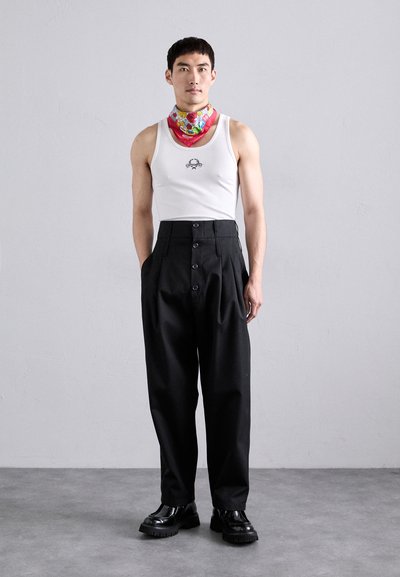 Hvid tanktop med et lille grafisk motiv, parret med højtaljede sorte bukser med fem knapper, og et farverigt mønstret tørklæde om halsen. Sorte sko.