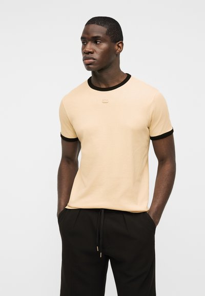 Νέος μαύρος άνδρας που φοράει μπεζ κοντομάνικη μπλούζα με μαύρο περίγραμμα και μαύρο παντελόνι, στέκεται με τα χέρια στις τσέπες, κοιτώντας προς τα αριστερά.