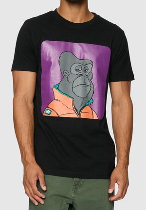 Uomo che indossa una maglietta nera con un gorilla cartone animato con giacca arancione su sfondo quadrato viola sul davanti.