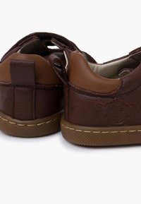 Scarpe in pelle marrone con superficie texture, dotate di suola beige e un emblema a faccina sorridente sul lato. Inclusa una linguetta sul tallone per indossarle facilmente.