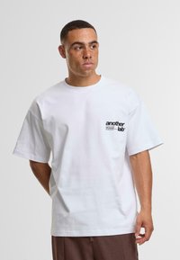 Tricou alb din bumbac cu mâneci scurte, având un logo imprimat negru pe pieptul stâng. Croială dreaptă cu un gât rotund.