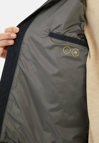 Herrenjacke mit beiger Außenseite und grauem Innenfutter, ausgestattet mit einer Brusttasche und wasserabweisenden, schmutzabweisenden Markierungen.