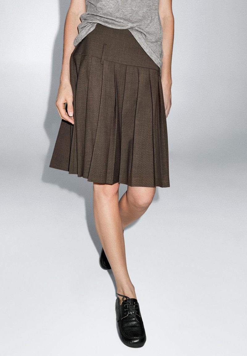 Femme portant une jupe plissée marron, une chemise grise rentrée et des chaussures noires à lacets, debout devant un fond clair uni.