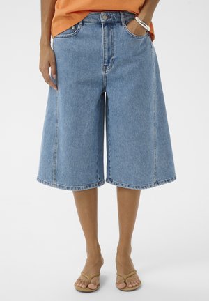 Persona que lleva pantalones cortos de mezclilla azul claro, sueltos y de longitud media, camiseta naranja, pulsera plateada y sandalias beige.