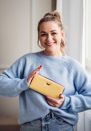 Yellow wallet with embossed dots and a logo, secured with a dark zipper. Held against a light blue sweater and denim jeans.