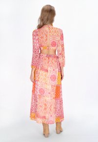 Langes, fließendes Kleid mit einem bunten Patchwork aus rosa, orange und gelben Mustern. Durchsichtiger Stoff mit einem Rückenausschnitt und langen Ärmeln.