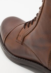 Gros plan d’une botte en cuir marron avec des lacets marron foncé et des coutures visibles sur une semelle en caoutchouc noire.