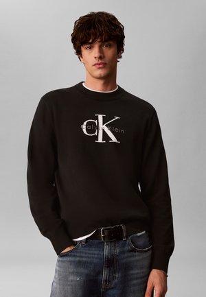 Jeune homme aux cheveux bouclés portant un pull noir Calvin Klein et un jean bleu, debout avec une main dans la poche sur fond gris.
