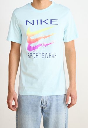 Mann trägt ein hellblaues Nike-T-Shirt mit bunten, verzerrten Swoosh-Logos und dem Schriftzug "SPORTSWEAR", kombiniert mit hellen Jeans.