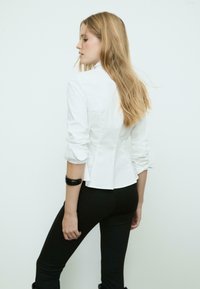 Weißes, tailliertes Blusenshirt mit hohem Kragen, langen Ärmeln und peplumförmigem Rücken. Kombiniert mit schwarzen, eng anliegenden Hosen. Glatte Textur, keine sichtbaren Muster.