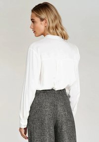 Biała jedwabna bluzka z długimi rękawami, z detalem w postaci plisy z tyłu. Noszona z szarymi spodniami o szerokich nogawkach z teksturowanego materiału. Prosty i klasyczny design.