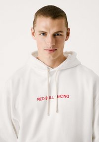 Sudadera blanca hecha de un tejido suave, con el texto "RED BULL RACING" en rojo. Diseño simple con cordones en la capucha. Textura suave.
