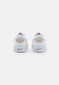 Bílé tenisky Nike s plochou podrážkou, jemným béžovým akcentem a vyraženým logem na patě, hladký kožený svršek, klasický design.