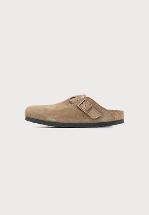 Sabot marron en daim à enfiler avec bride à boucle réglable et semelle noire sur fond clair neutre.