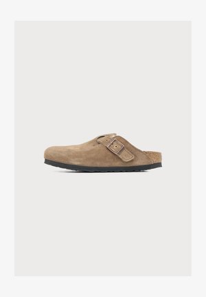 Sabot marron en daim à enfiler avec bride à boucle réglable et semelle noire sur fond clair neutre.