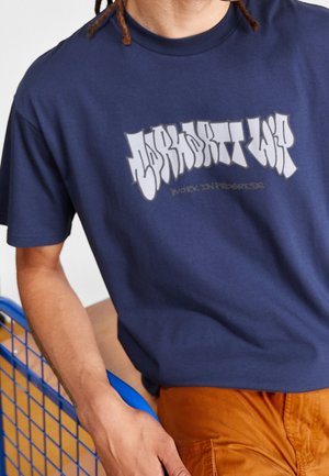 Osoba ubrana w granatowy t-shirt z białym napisem graffiti i pomarańczowe spodnie, opierająca rękę na niebieskiej metalowej poręczy.
