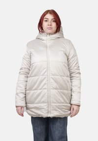 Cappotto imbottito beige chiaro con cappuccio, pannelli trapuntati, cerniera frontale e polsini elasticizzati. Indossato sopra jeans blu.