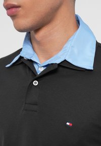 Detailný záber na osobu, ktorá má na sebe čiernu polo košeľu s dvoma gombíkmi a viditeľným malým obdĺžnikovým logom, vrstvenou nad svetloblú košeľou s golierom.