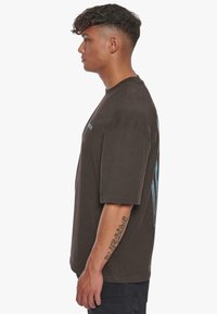 Brun oversized t-skjorte med rund hals og et grafisk design på baksiden. Korte ermer og en avslappet passform.