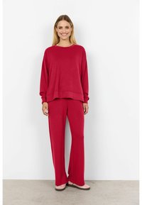 Top roșu, cu mâneci lungi și croială lejeră, asortat cu pantaloni largi de aceeași culoare. Textură moale, design casual, purtat cu încălțăminte roz deschis.