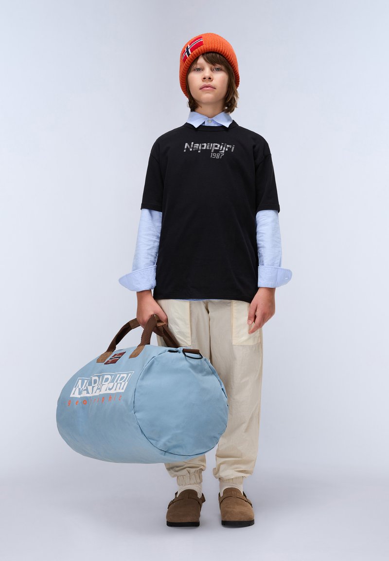 Ljusblå duffelväska med bruna handtag, rund form, och logotyp i vitt och rött. Modell som bär en svart t-shirt, beige byxor och en orange mössa.