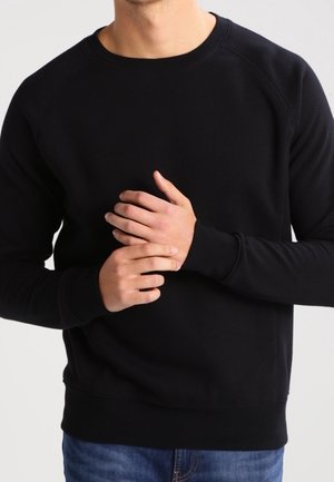 Mężczyzna w jednolitym czarnym swetrze z długim rękawem i niebieskich dżinsach, ręce złączone na poziomie klatki piersiowej na jednolitym tle.