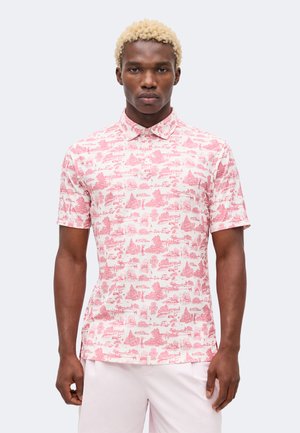 Man met kort blond haar, die een roze-wit patroon korte mouwen overhemd met knopen en lichte broek draagt.