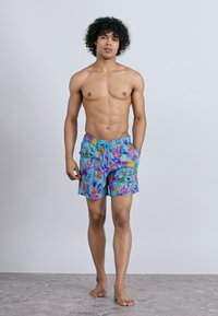 Tânăr cu păr creț, purtând șorturi de baie florale albastre, stând desculț pe un podea gri, pe un fundal simplu de culoare deschisă.