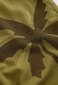 Olivengrønt stof med et stort, tekstureret brunt bladmotiv med lysere brune accenter, der fremviser en blanding af glatte og broderede teksturer.