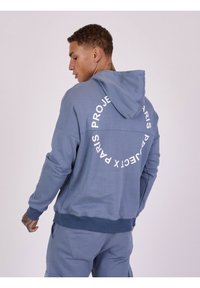 Lichtblauwe hoodie met een trekkoord en een ronde witte print op de achterkant, gemaakt van zacht materiaal. Heeft ribbelboorden en een ribbelzoom.