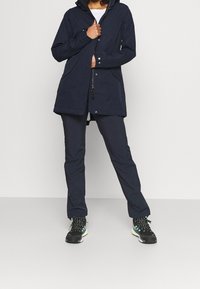 Chaqueta impermeable de color marino con capucha, cierre de cremallera y dos bolsillos frontales, combinada con pantalones a juego y zapatillas negras con acentos verdes.